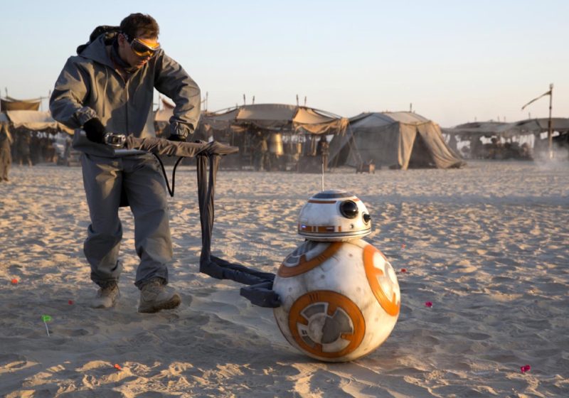 Puppeteer Brian Herring, dressed in a grey greenscreen suit and goggles, operates the BB-8 droid puppet across a sandy desert set during the filming of the Star Wars sequel trilogy.