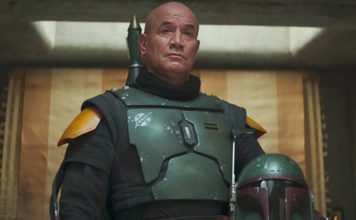 A medium shot of Temuera Morrison as Boba Fett, wearing his green and black Beskar armor without a helmet, standing in his palace on Tatooine.
