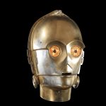 A screen-matched C-3PO prop head from Star Wars: The Empire Strikes Back with glowing yellow eyes, showing authentic weathering and gold-and-silver plating against a black background.