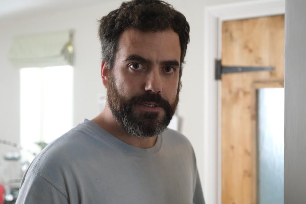 Actor Daniel Ings looking directly at the camera with a serious expression and a full beard, wearing a light blue t-shirt in a domestic setting.
