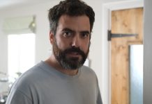 Actor Daniel Ings looking directly at the camera with a serious expression and a full beard, wearing a light blue t-shirt in a domestic setting.