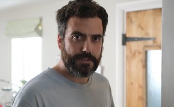 Actor Daniel Ings looking directly at the camera with a serious expression and a full beard, wearing a light blue t-shirt in a domestic setting.