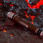 A close-up view from above of Jedi Devon Izara's lightsaber hilt with a brown textured grip and silver accents, resting on a field of dark, cracked volcanic rock with glowing red lava visible in the fissures.