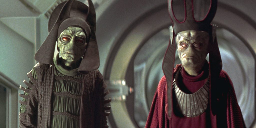 Two Neimoidian characters, Nute Gunray and Rune Haako from the Star Wars prequel trilogy, wearing ornate Trade Federation robes and tall ceremonial headdresses while standing inside a sterile, metallic corridor.