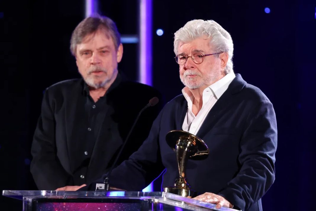George Lucas holds a gold Saturn Award at a podium while Mark Hamill stands behind him on stage.