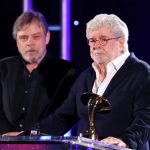 George Lucas holds a gold Saturn Award at a podium while Mark Hamill stands behind him on stage.