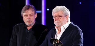 George Lucas holds a gold Saturn Award at a podium while Mark Hamill stands behind him on stage.