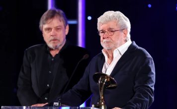 George Lucas holds a gold Saturn Award at a podium while Mark Hamill stands behind him on stage.