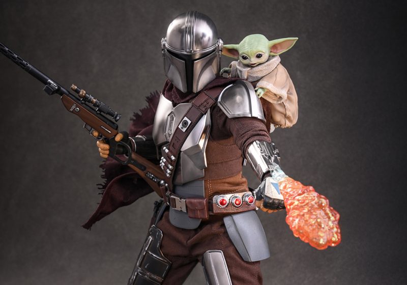 A highly detailed 1/6th scale Hot Toys action figure of The Mandalorian in silver Beskar armor, holding a sniper rifle and firing a flamethrower from his gauntlet, with a small Grogu figure magnetically attached to his shoulder.