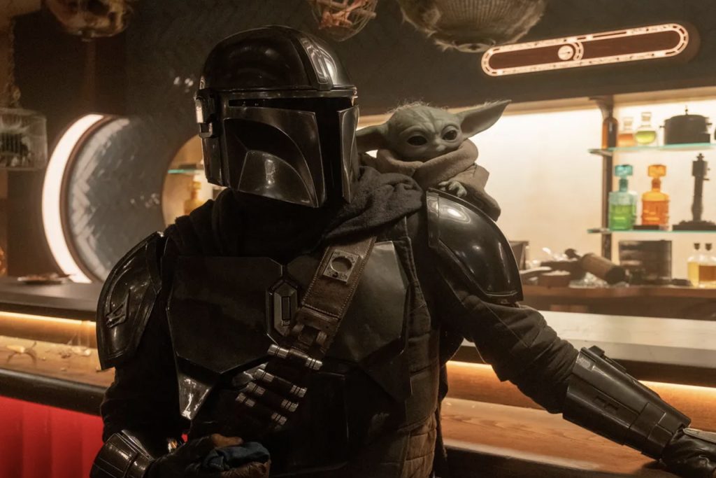 The Mandalorian wearing his shiny silver Beskar armor and helmet with the small, green, large-eared Grogu sitting on his right shoulder inside a dimly lit cantina.