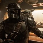 The Mandalorian wearing his shiny silver Beskar armor and helmet with the small, green, large-eared Grogu sitting on his right shoulder inside a dimly lit cantina.