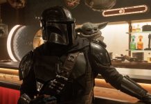 The Mandalorian and Grogu Runtime: How Long is the New Star Wars Movie? The Mandalorian wearing his shiny silver Beskar armor and helmet with the small, green, large-eared Grogu sitting on his right shoulder inside a dimly lit cantina.
