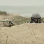 Grogu using a telescope while the Mandalorian looks on from behind a sand dune.