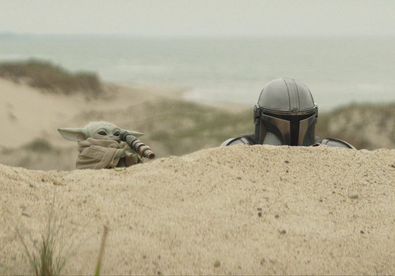 Grogu using a telescope while the Mandalorian looks on from behind a sand dune.