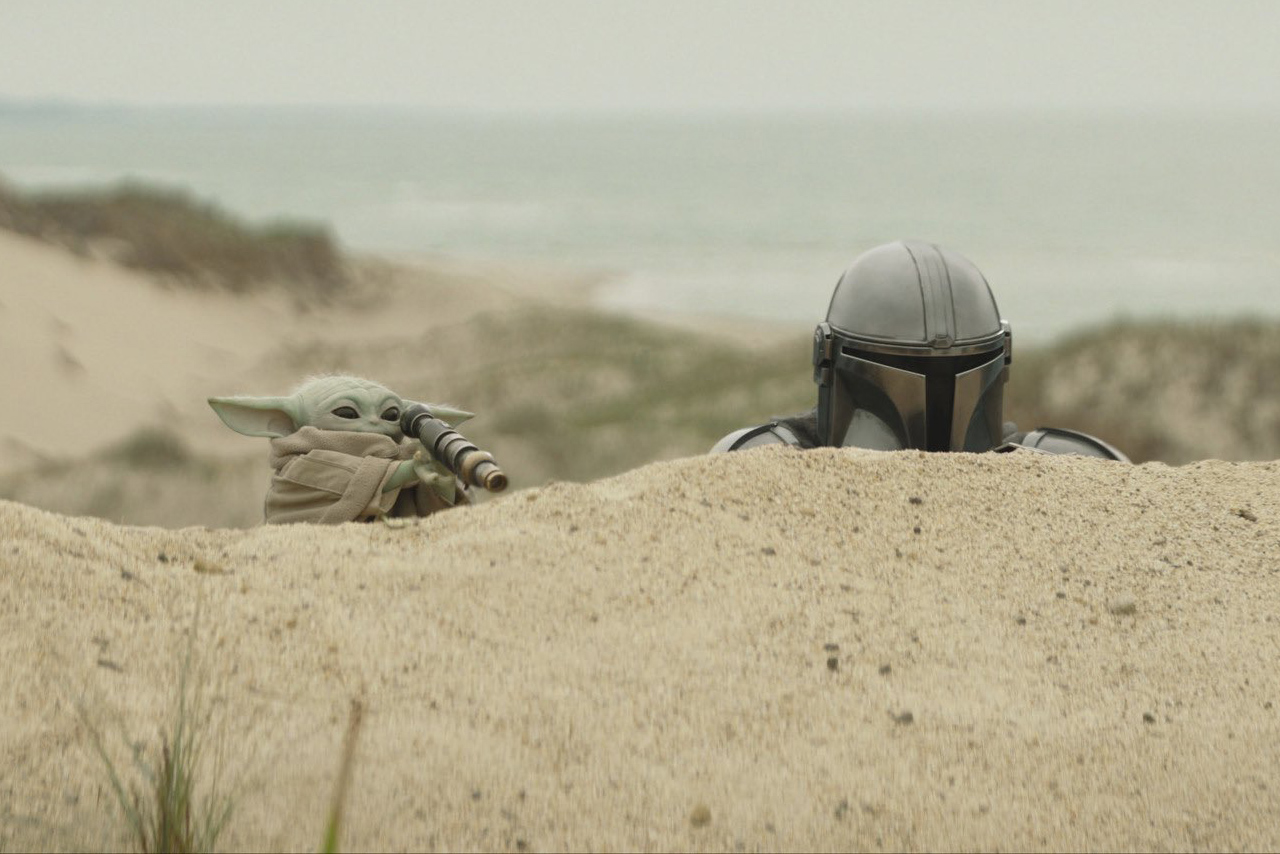 Grogu using a telescope while the Mandalorian looks on from behind a sand dune.