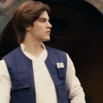 A face character performer portraying a young Han Solo in a blue vest and white tunic standing in front of a Black Spire Outpost doorway at Star Wars: Galaxy's Edge.