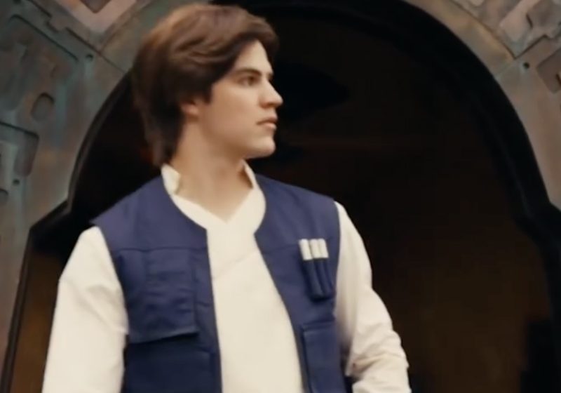 A face character performer portraying a young Han Solo in a blue vest and white tunic standing in front of a Black Spire Outpost doorway at Star Wars: Galaxy's Edge.