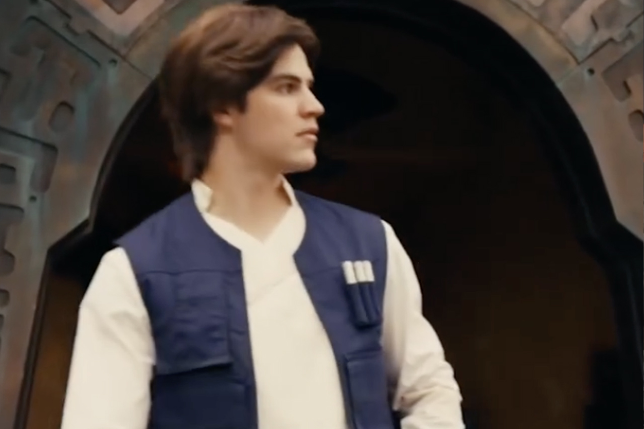 A face character performer portraying a young Han Solo in a blue vest and white tunic standing in front of a Black Spire Outpost doorway at Star Wars: Galaxy's Edge.