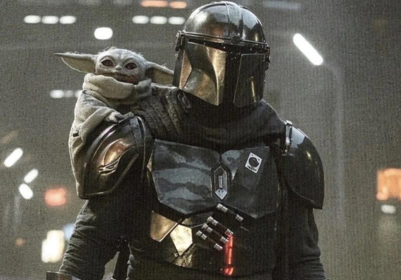 The Mandalorian standing in a dark, industrial setting with Grogu (Baby Yoda) perched on his shoulder, both looking forward.