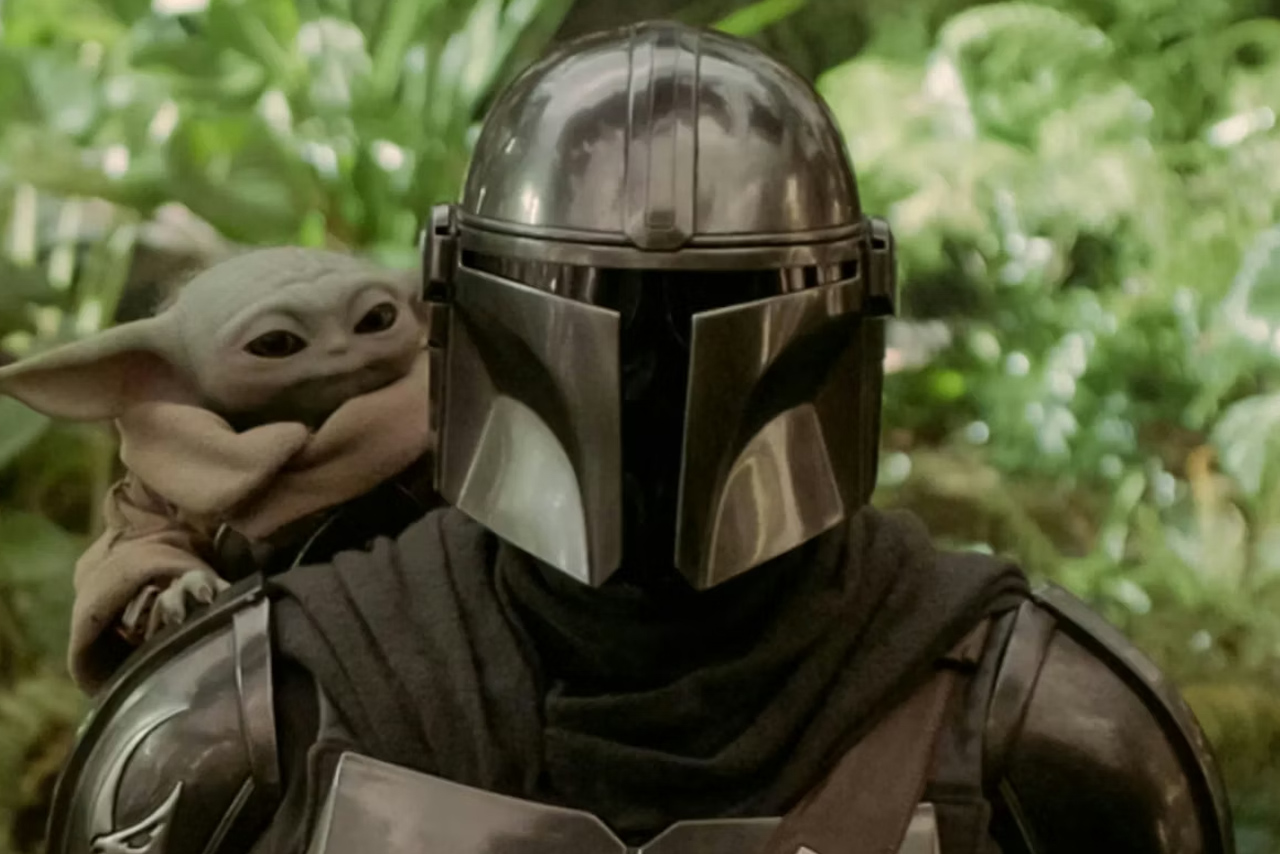 Din Djarin in full beskar armor with Grogu looking over his right shoulder in a lush green jungle environment from the Star Wars film.